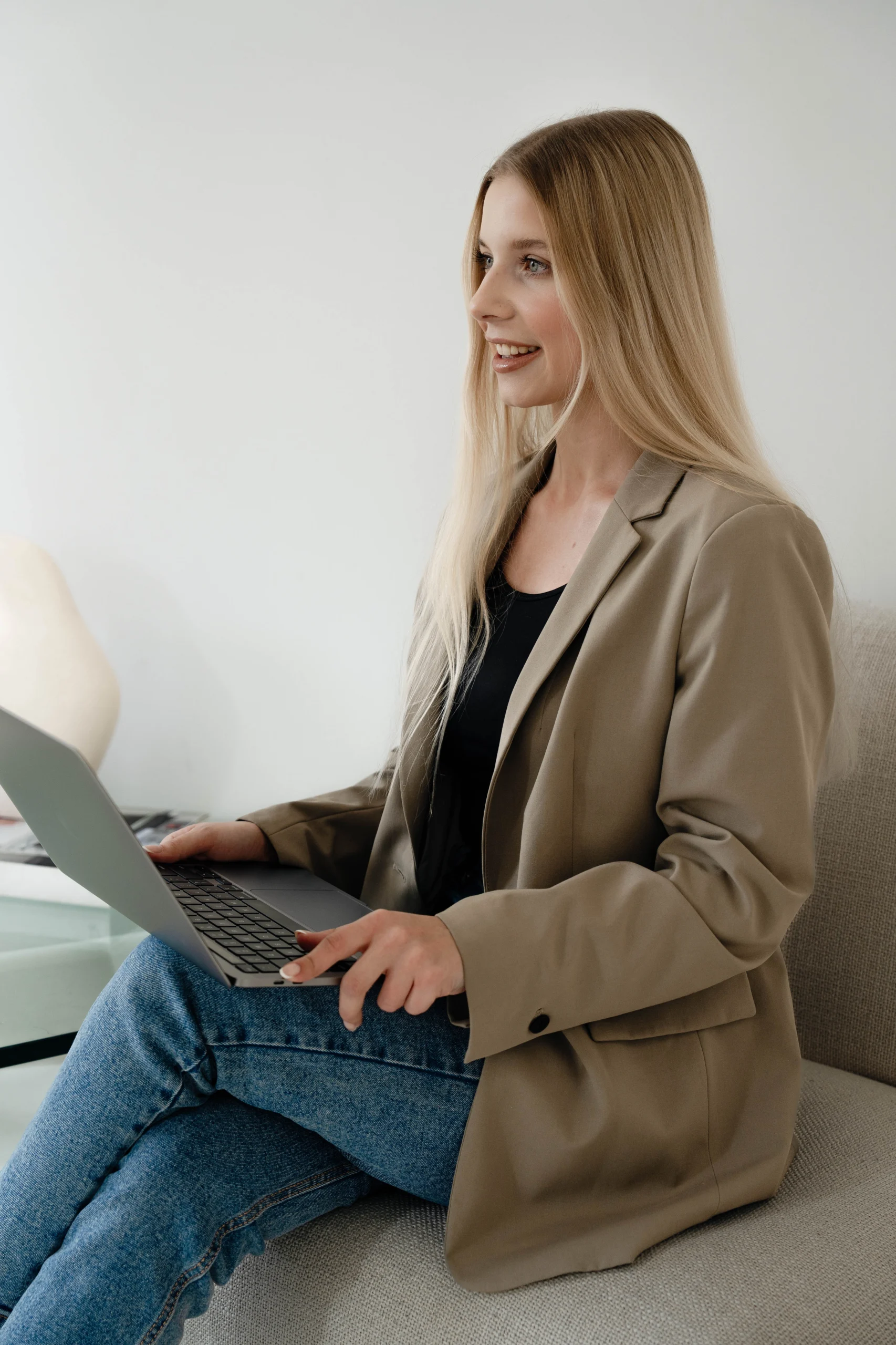 Młoda kobieta o blond włosach siedzi na kanapie i trzyma laptop na kolanach. Jest ubrana w dżinsy i beżową marynarkę.